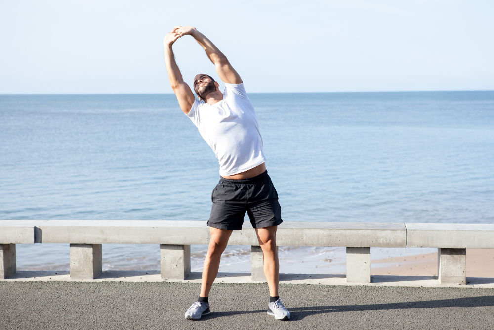 Man performing a standing side stretch by the seaside, raising arms overhead to stretch the torso during an outdoor workout.